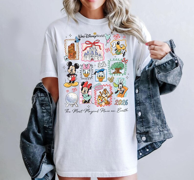Może przedstawiać: Biała koszulka z kolorowymi postaciami Disneya oraz napisami "Walt Disney World" i "2026". Koszulka zawiera r&oacute;wnież frazę "The Most Magical Place on Earth".