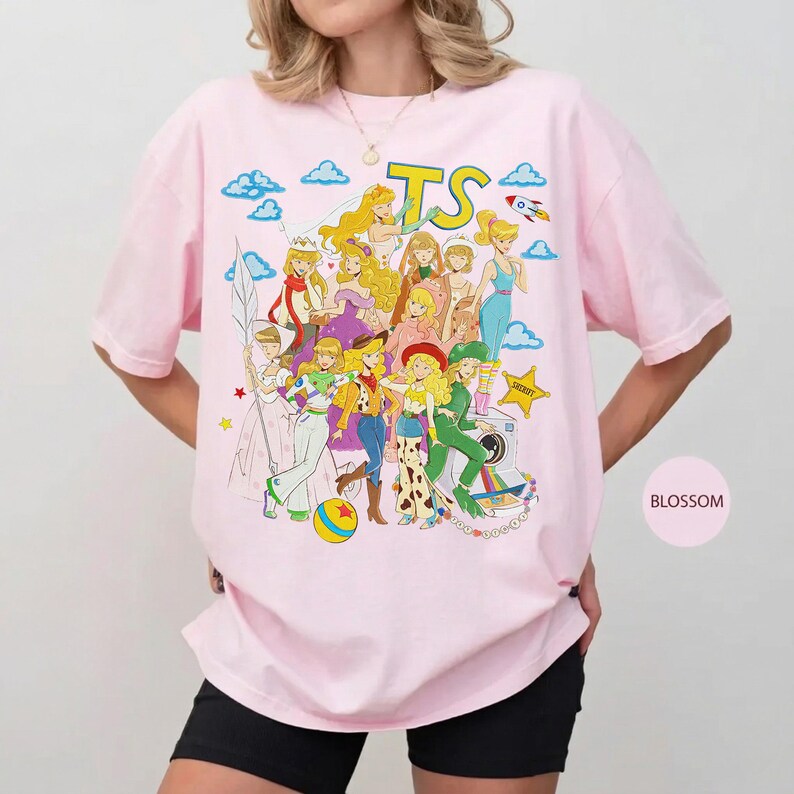 Puede incluir: Una camiseta de manga corta de color rosa claro con un gran estampado gr&aacute;fico de varios personajes animados con diversos atuendos. El texto TS est&aacute; impreso en amarillo en la parte superior. El dise&ntilde;o incluye nubes, un cohete y una estrella.