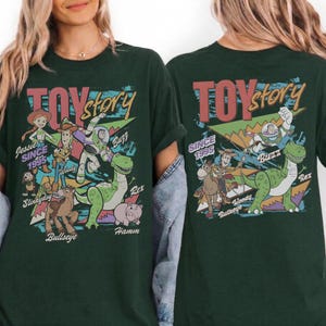 May include: A dark green t-shirt featuring a graphic print of Toy Story characters including Jessie, Woody, Buzz Lightyear, Slinky Dog, Bullseye, Rex, and Hamm. The design includes the text Toy Story and Since 1995 with colorful geometric shapes.