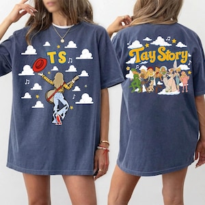 May include: A navy blue t-shirt featuring a graphic print. The front shows a person with a guitar, a red hat, and the text TS against a cloud background. The back displays a group of figures and the text Tay Story with clouds and stars.