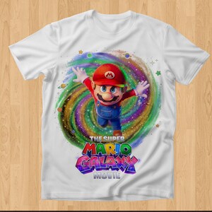 May include: A white short-sleeve t-shirt featuring a graphic of Mario in a red cap and blue overalls leaping in front of a colorful swirling galaxy background with small planets and stars, with the text THE SUPER MARIO GALAXY MOVIE below.