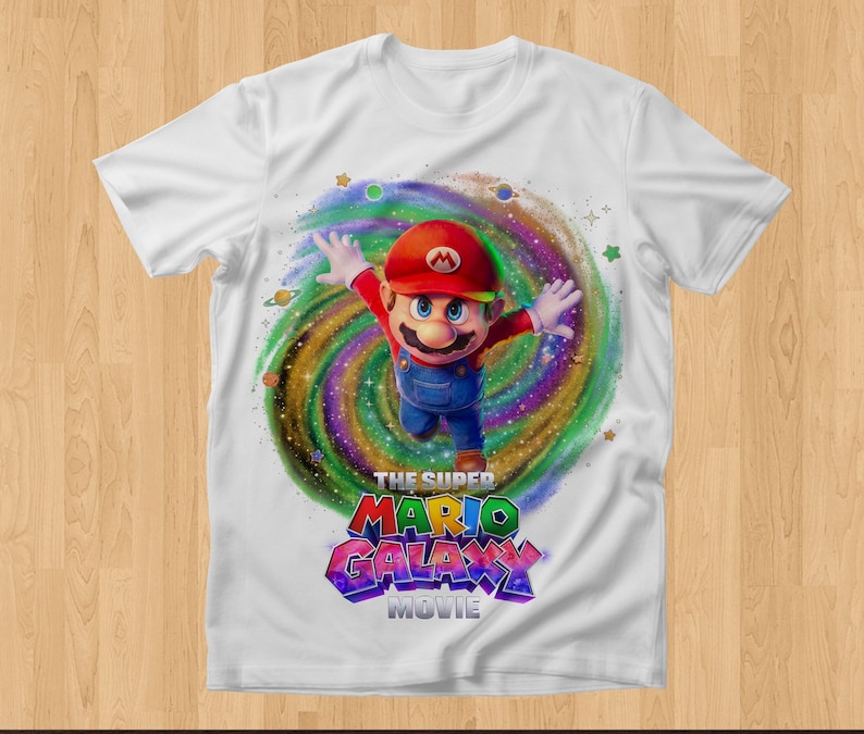 May include: A white short-sleeve t-shirt featuring a graphic of Mario in a red cap and blue overalls leaping in front of a colorful swirling galaxy background with small planets and stars, with the text THE SUPER MARIO GALAXY MOVIE below.