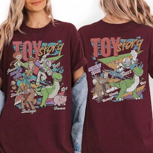 May include: A maroon short-sleeve t-shirt featuring a graphic print of Toy Story characters including Jessie, Woody, Buzz Lightyear, Rex, Slinky Dog, Bullseye, and Hamm. The text reads Toy Story and Since 1995, with character names printed below.
