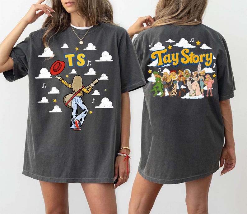 May include: A charcoal gray short-sleeve t-shirt featuring a graphic print. The front displays a figure with a guitar, a red hat, and the text TS. The back shows a group of illustrated characters and the text Tay Story, both set against clouds.