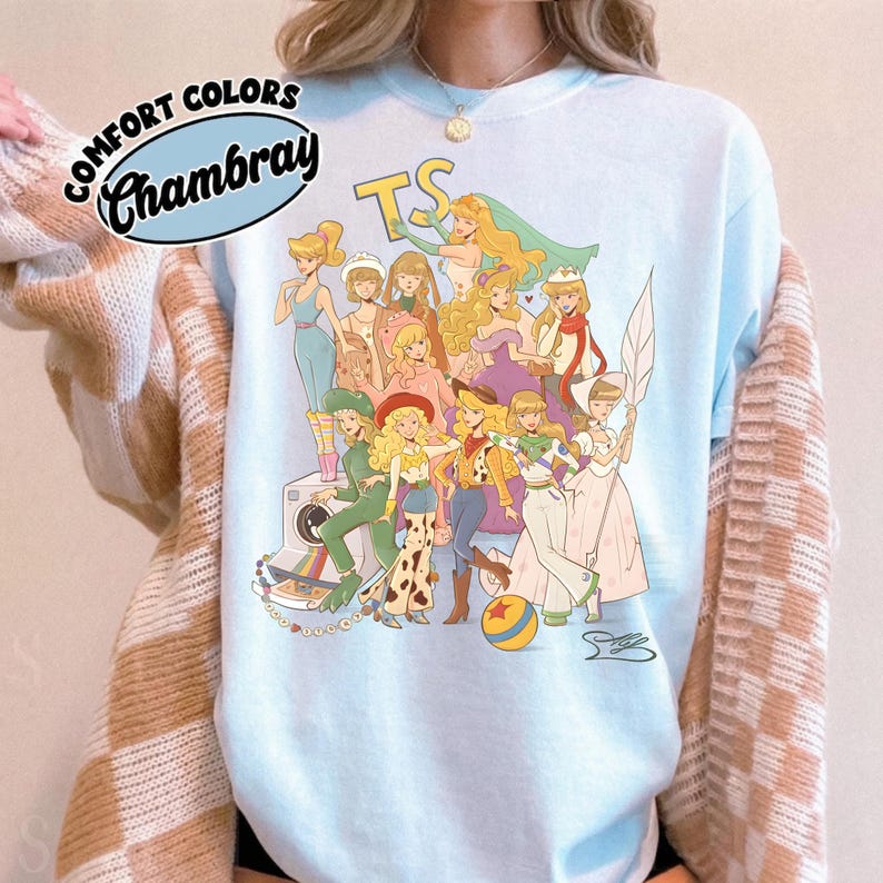 Puede incluir: Una camiseta de cuello redondo azul claro con un estampado gr&aacute;fico de varios personajes animados con diversos atuendos, incluyendo un sombrero de vaquero, un traje espacial y un vestido de gala. El texto "COMFORT COLORS" y "Chambray" aparece arriba.