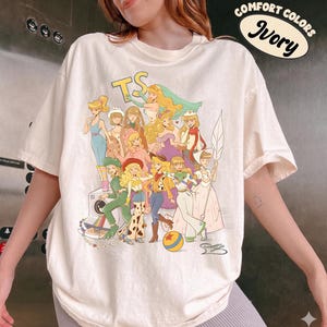 Puede incluir: Una camiseta de manga corta color marfil con un estampado gr&aacute;fico de varios personajes animados con diversos atuendos. El texto "COMFORT COLORS Ivory" y "TS" es visible. El dise&ntilde;o incluye una pelota colorida y una ilustraci&oacute;n de una c&aacute;mara.
