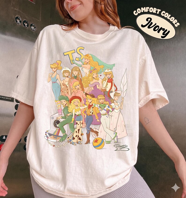 Puede incluir: Una camiseta de manga corta color marfil con un estampado gr&aacute;fico de varios personajes animados con diversos atuendos. El texto "COMFORT COLORS Ivory" y "TS" es visible. El dise&ntilde;o incluye una pelota colorida y una ilustraci&oacute;n de una c&aacute;mara.