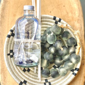 May include: A clear glass diffuser bottle with the text "Tiny Bandit Crystal Diffuser" sits in a woven basket with black and white accents. The basket holds green glass beads. Two white diffuser sticks are placed next to the bottle.