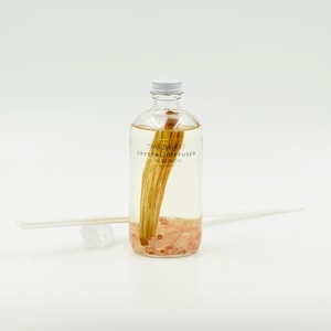 May include: A clear glass bottle with a white cap, containing a light-colored liquid, a wooden stick, and pink crystals. The bottle has the text "TINY BANDIT CRYSTAL DIFFUSER" printed on it. Two white diffuser sticks and a small crystal are next to the bottle.