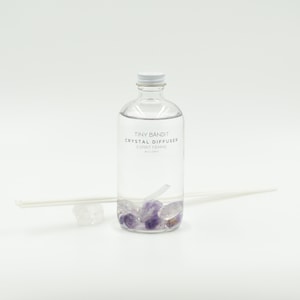May include: A clear glass bottle labeled "TINY BANDIT CRYSTAL DIFFUSER ESPRIT FEMME" with a white cap, filled with liquid and purple crystals. Two white diffuser sticks and a clear crystal are next to the bottle. The background is white.