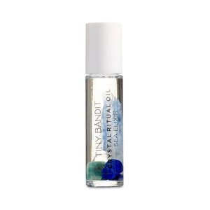 Op de afbeelding: Een heldere glazen rollerfles met een witte dop. De fles bevat een heldere vloeistof en kleine blauwe en groene stenen. De tekst "TINY BANDIT CRYSTAL RITUAL OIL SEA ELIXIR" staat op de fles.
