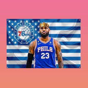 May include: A rectangular wall flag featuring a basketball player wearing a blue Philadelphia 76ers jersey with the number 23 and a gold crown on their head. The background displays a blue and white striped flag with a circular 76ers team logo.