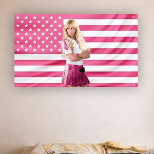 May include: A framed print featuring a person standing in front of a pink and white American flag. The person is wearing a white top, a pink skirt, and has long blonde hair. The flag design is a vibrant pink and white color scheme.