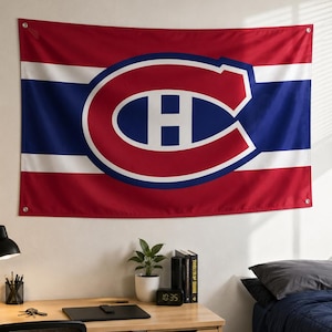 Op de afbeelding: Een rood, wit en blauw vlag met het logo van de Montreal Canadiens. Het logo is een grote, gestileerde "C" met een hockeypuck-vorm erin en de letter "H" in het midden. De vlag hangt aan een witte muur.