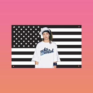 Peut inclure: Un t-shirt blanc avec le texte "ecko unltd" en bleu et noir, porté avec une casquette blanche et des bracelets. Le fond présente un motif de drapeau américain noir et blanc.