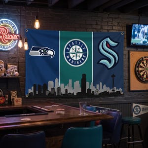 Puede incluir: Un estandarte de pared rectangular con tem&aacute;tica deportiva de Seattle sobre un fondo azul oscuro y verde azulado. Muestra el logotipo de los Seattle Seahawks, los Seattle Mariners y los Seattle Kraken sobre una silueta del horizonte de Seattle.