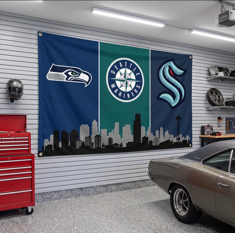 Puede incluir: Una gran bandera de pared rectangular con un dise&ntilde;o tricolor vertical en azul marino y verde azulado. La bandera muestra el logotipo de los Seattle Seahawks, el de los Seattle Mariners y el de los Seattle Kraken sobre una silueta negra.