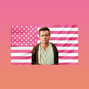 May include: A rectangular flag with a pink American flag design. The flag has white stars and pink stripes. A person wearing a brown robe and cream-coloured tunic is positioned in front of the flag.