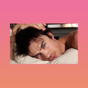 May include: A person with dark hair and blue eyes is lying on a white pillow, looking towards the viewer. The person's arm is resting on their head. The background is a gradient of pink and orange.