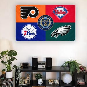 May include: A rectangular wall flag featuring four quadrants in orange, red, blue, and dark green. It displays the Philadelphia Flyers, Phillies, 76ers, and Eagles logos, with the Philadelphia Union crest centered in the middle of the flag.