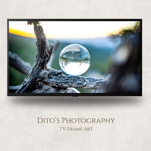 May include: A photograph displayed on a TV screen. The image shows a clear glass sphere on a dark brown, weathered tree branch. The sphere reflects a landscape with a sunset and trees. The text "DITO'S PHOTOGRAPHY TV FRAME ART" is below the TV.