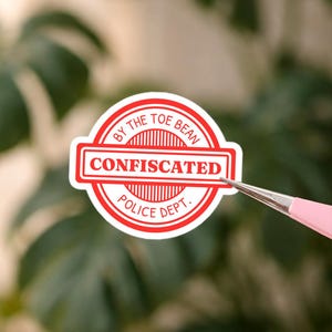May include: A red and white sticker with the text "CONFISCATED" in a rectangular box, surrounded by a circular border that reads "BY THE TOE BEAN POLICE DEPT." The sticker is held by pink tweezers against a blurred green backdrop.