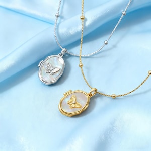 Op de afbeelding: Twee kettingen met vlinderhangers. De ene ketting is zilverkleurig, de andere goudkleurig. Beide hangers hebben een parelmoeren achtergrond en een vlinderontwerp. De kettingen liggen op een lichtblauwe satijnen achtergrond.