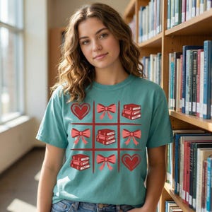 May include: Teal t-shirt featuring a tic-tac-toe design with red hearts, bows, and stacks of books. The shirt is made of a soft material and is suitable for casual wear. The design is centered on the front of the shirt.