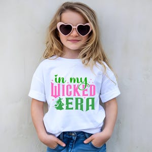 May include: A white t-shirt with the text "in my WICKED ERA" in pink and green, featuring a witch graphic. The shirt is worn by a child wearing pink heart-shaped sunglasses.