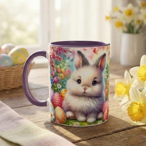 May include: A ceramic mug with a purple handle and interior. The mug features a colorful illustration of a fluffy white bunny surrounded by Easter eggs and flowers. The mug is on a wooden surface.