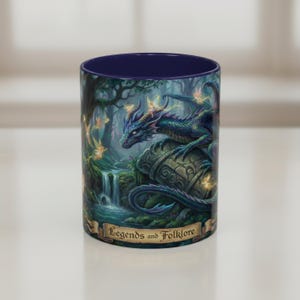 May include: A cylindrical mug with a dark blue interior and a fantasy scene. The exterior features a blue and green dragon perched on a stone, with fairies, a waterfall, and the text "Legends and Folklore".