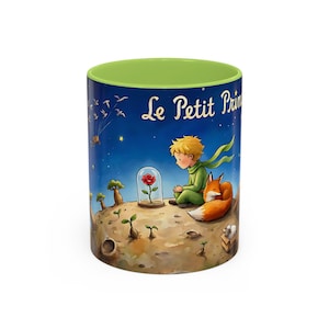 May include: A ceramic mug featuring a scene from "Le Petit Prince." The design shows the Little Prince, a rose in a glass dome, and a fox. The mug has a green interior and a blue exterior with the title in white.