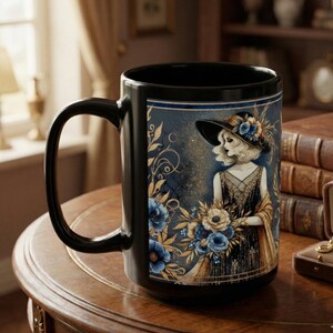 May include: Black ceramic mug featuring a vintage illustration of a woman in a hat holding flowers. The design includes blue and gold floral accents against a dark blue background. The mug is on a wooden surface.