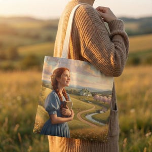May include: A cream-coloured tote bag displaying a scenic illustration of a woman in a blue dress holding a book. The artwork shows a winding road, a white house, and a river under a cloudy sky. The bag has a white canvas strap.