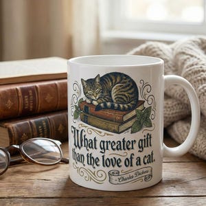 Może przedstawiać: Biały ceramiczny kubek z ilustracją śpiącego kota pręgowanego na stosie książek. Kubek zawiera cytat "What greater gift than the love of a cat" i jest ustawiony na drewnianej powierzchni z książkami i okularami.