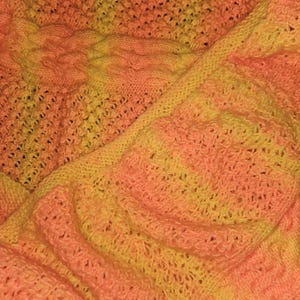 May include: Close-up of a hand-knitted blanket in shades of orange and yellow. The blanket features various textured patterns, including cable knit and openwork designs. The color gradient creates a warm, inviting aesthetic.