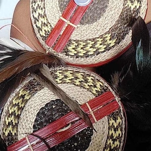 Puede incluir: Dos sombreros tejidos con un diseño circular en tonos marrón, negro y amarillo. Cada sombrero presenta un acento rojo y un botón. Plumas están unidas a los sombreros, agregando un elemento decorativo.