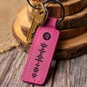 May include: A pink leather keychain with a Spotify code and a key ring. The keychain is rectangular with a stitched border and a black Spotify logo. A gold-colored key is attached to the ring.