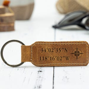 May include: A brown leather keychain with a metal ring. The keychain has the coordinates 34°02'35"N 118°16'02"W and a compass rose design. The leather is stitched around the edges.