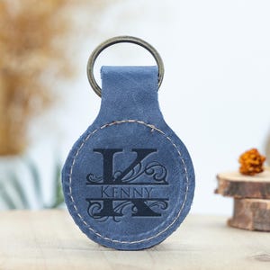 May include: A blue leather keychain with a metal ring. The keychain features the letter "K" and the name "Kenny" engraved in black. The keychain is oval-shaped with a stitched border.