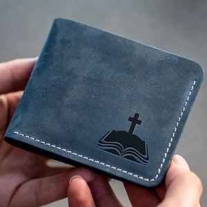 May include: A blue leather wallet with a white stitched border. The wallet features a black graphic of an open book with a cross on top. The wallet is held in a person's hands.