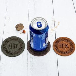 May include: Three circular coasters with a Greek key border pattern. One dark gray coaster features the monogram JHT, one brown coaster features the monogram HK, and a blue Bud Light aluminum beverage can sits on the center dark brown coaster.