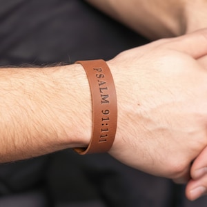 May include: A brown leather bracelet with the text "PSALM 91:11" engraved on it. The bracelet is worn on a wrist, showcasing its simple design and religious inscription. The leather appears to be a medium brown color.