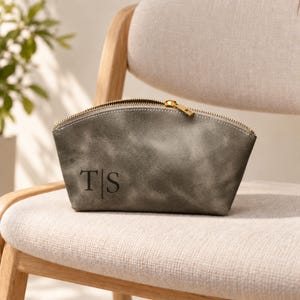 May include: A gray, semi-circular zippered pouch with a distressed texture. The front features the black, embossed letters T and S separated by a vertical line. The pouch has a gold-toned metal zipper closure along the top edge.