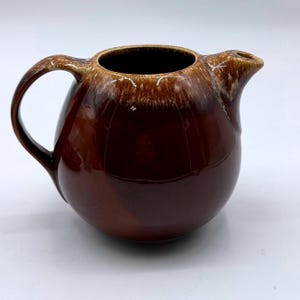 May include: A round ceramic pitcher with a glossy dark brown glaze. The upper rim features a lighter tan and cream-colored drip pattern that cascades down from the opening. It has a curved handle on the left side and a small pouring spout on the right.