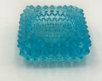 Könnte beinhalten: Eine quadratische, hellblaue Glasschale mit einem Noppenmuster, bei dem erhabene, spitze Noppen die Außen- und Innenflächen bedecken. Das durchscheinende Glas hat eine gleichmäßige hellblaue Farbe und ein markantes, geometrisches Design.