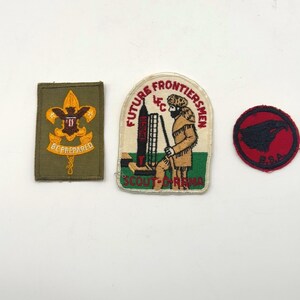 May include: Three embroidered patches: a rectangular olive green patch with a yellow eagle emblem and the text BE PREPARED, a shield-shaped patch depicting a person in frontier clothing with a rocket and the text FUTURE FRONTIERSMEN SCOUT-O-RAMA, and a circular red patch with a black eagle head and the text B.S.A.