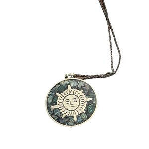 May include: A circular pendant necklace featuring a silver-toned sun face emblem centered on a mosaic background of dark green and blue stone fragments. The pendant hangs from a thin, dark brown cord, showcasing a detailed sun design with rays.