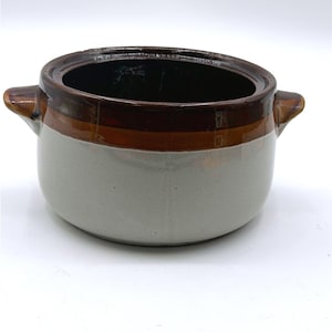 May include: A round ceramic pot with two small side handles. The exterior features a two-tone design with a light gray speckled base and a wide, dark brown glazed band around the upper rim. The interior of the pot is finished in a dark brown glaze.