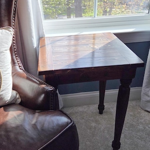May include: A dark brown wooden side table with a rectangular top and four turned legs. The table is positioned next to a brown leather armchair and a window with a view of trees. The table's top has a slatted design.